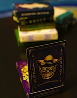 A playing card featuring a stylized sugar skull design, labeled 'Joker', is prominently displayed in the foreground. The card has an intricate design with a hat on the skull and decorative elements in gold. In the background, multiple stacks of board games or boxes are slightly out of focus, placed on a dark tabletop.