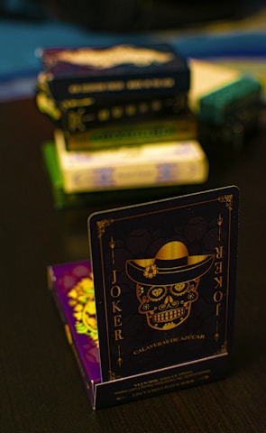 A playing card featuring a stylized sugar skull design, labeled 'Joker', is prominently displayed in the foreground. The card has an intricate design with a hat on the skull and decorative elements in gold. In the background, multiple stacks of board games or boxes are slightly out of focus, placed on a dark tabletop.