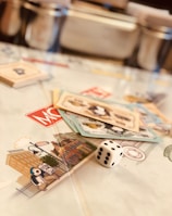 A tabletop setup featuring Monopoly-themed cards and a single die placed on a game board. The cards display various illustrations, while the die shows the number five. The scene is softly lit, creating a warm and inviting atmosphere.
