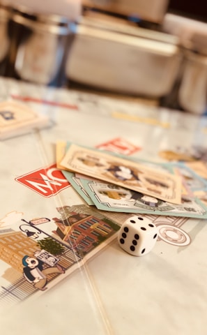 A tabletop setup featuring Monopoly-themed cards and a single die placed on a game board. The cards display various illustrations, while the die shows the number five. The scene is softly lit, creating a warm and inviting atmosphere.