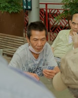 Several individuals gather outdoors, sitting on benches and engaging in a game of cards. The focal point is on a man holding a fan of playing cards, with others observing or participating. Potted plants and a red railing are visible in the background, contributing to the setting's casual and social atmosphere.