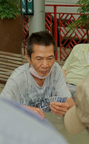 Several individuals gather outdoors, sitting on benches and engaging in a game of cards. The focal point is on a man holding a fan of playing cards, with others observing or participating. Potted plants and a red railing are visible in the background, contributing to the setting's casual and social atmosphere.