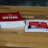 A wooden table features a stack of cards with a red cover labeled 'Exploding Kittens'. Next to it, a single card is laid out showing artwork and text related to the game. The setting is casual and likely related to a tabletop gaming session.