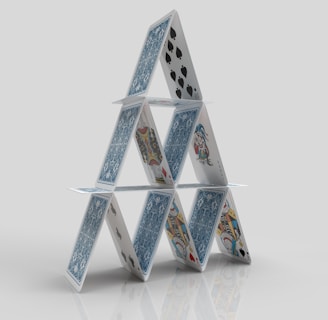 A structure composed of playing cards arranged in a triangular formation, resembling a house of cards. The cards feature various designs and suits, including spades and face cards, standing on a reflective surface against a plain background.