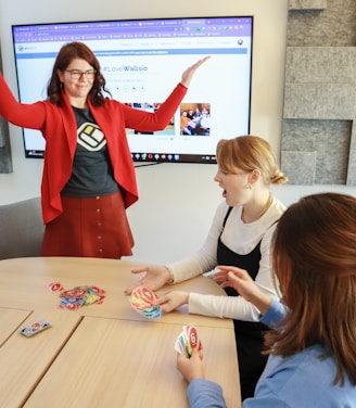 Four people are engaged in a lively game around a wooden table. One person stands with raised arms, while three others are seated, focusing on colorful cards in their hands. There is a computer monitor in the background displaying a webpage, and the room features a modern design with gray acoustic panels on the walls.