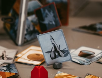 A tabletop game setup with various illustrated cards displayed on a surface. A red, geometric game piece is positioned next to the cards, adding a pop of color. The cards have images and symbols, suggesting a fantasy or strategy theme. The background shows a partially obscured game box with text.