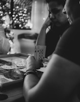 Three people are engaged in a card game, sitting around a table in a cozy, dimly lit setting. One player is holding an Ace and a Ten of diamonds. There are fairy lights and a blurred Christmas tree in the background, adding a festive and warm atmosphere to the scene.