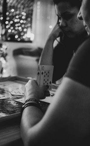 Three people are engaged in a card game, sitting around a table in a cozy, dimly lit setting. One player is holding an Ace and a Ten of diamonds. There are fairy lights and a blurred Christmas tree in the background, adding a festive and warm atmosphere to the scene.
